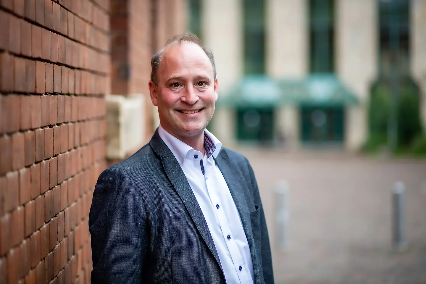 Ein Mann mit Anzug steht vor einer Backsteinmauer und lächelt in die Kamera.