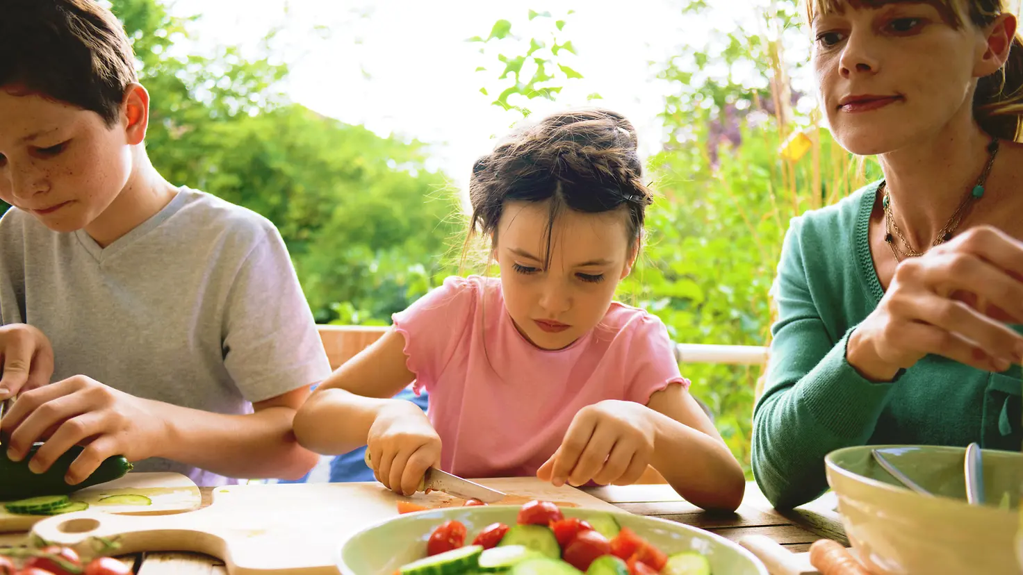 Eine Mutter und zwei Kinder sitzen an einem Tisch und schneiden Gemüse.