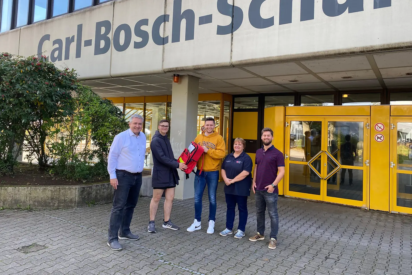 Drei Männer, ein Schüler und eine Frau stehen vor einer Schule. Der Schüler bekommt einen Erste-Hilfe-Rucksack überreicht.