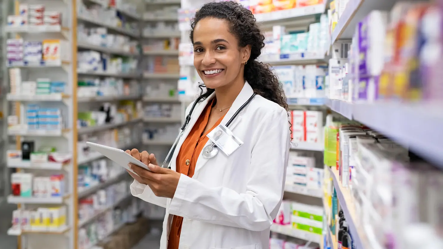 Eine Frau mit weißem Kittel und Stethoskop steht in einer Apotheke. Im Hintergrund sind Regale mit Medikamenten zu sehen.