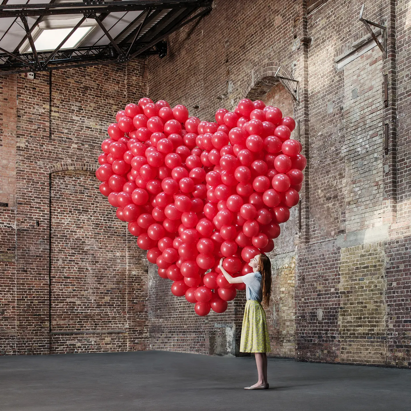 Junge Frau steht in einer Halle und umarmt ein grosses Herz aus roten Luftballons