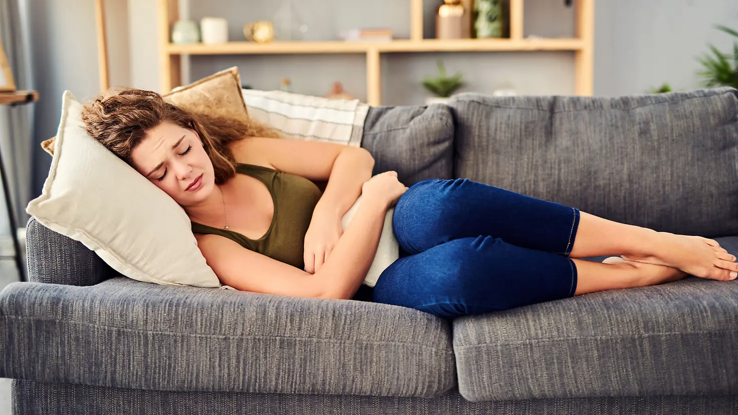 Eine junge Frau liegt mit Endometriose-Schmerzen und einer Wärmflasche auf dem Sofa.