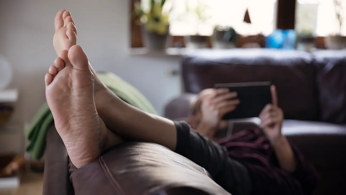 Eine Person liegt entspannt auf der Couch, hat die nackten Füße hochgelegt und liest ein Buch. Es kann der Prophylaxe gegen Fußpilz dienen, häufiger die Schuhe und Strümpfe auszuziehen.