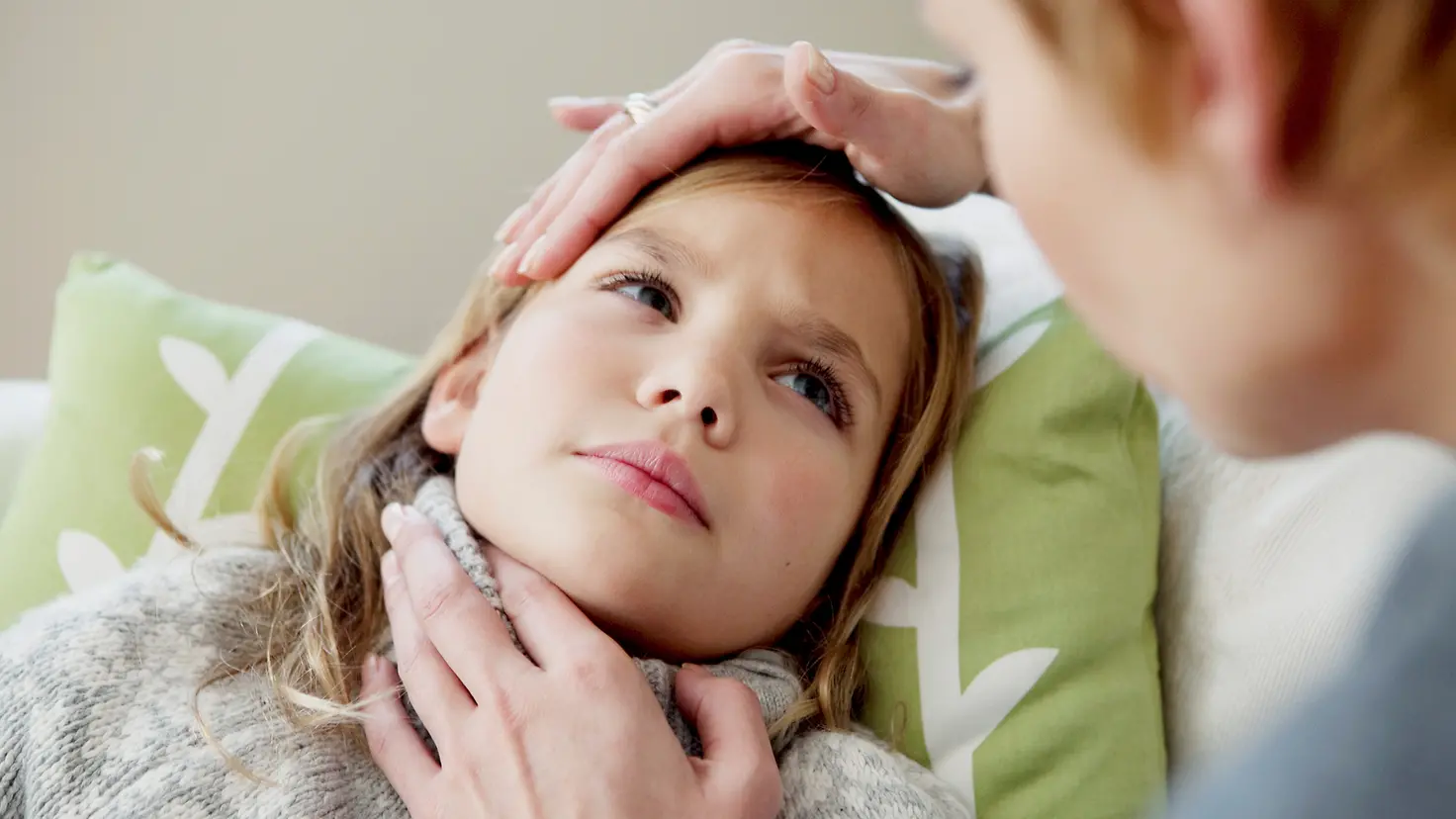 Ein Mädchen mit einer Tonsillitis liegt auf der Couch und ihre Mutter hat ihre Hand auf den Hals des Mädchens gelegt.