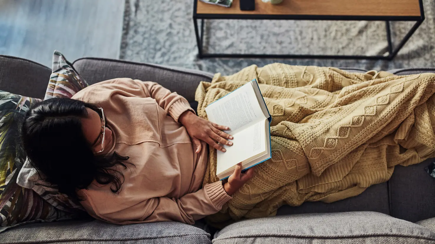 Eine Von-Oben-Aufnahme: eine Frau liegt zugedeckt auf einer Couch und liest in einem Buch.