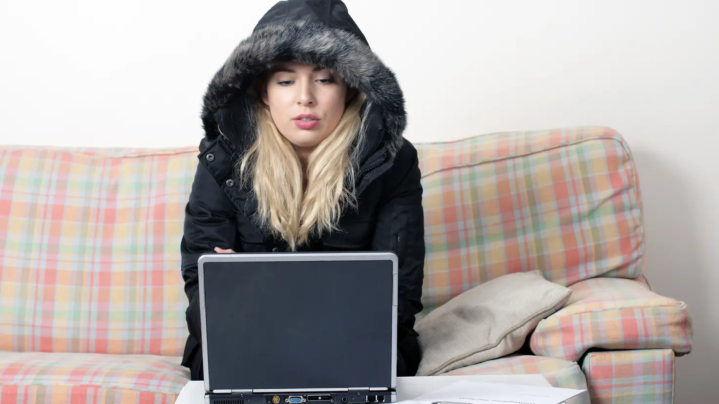 Eine Frau sitzt im waren Mantel in ihrer Wohnung auf einem Sofa vor ihrem Laptop.
