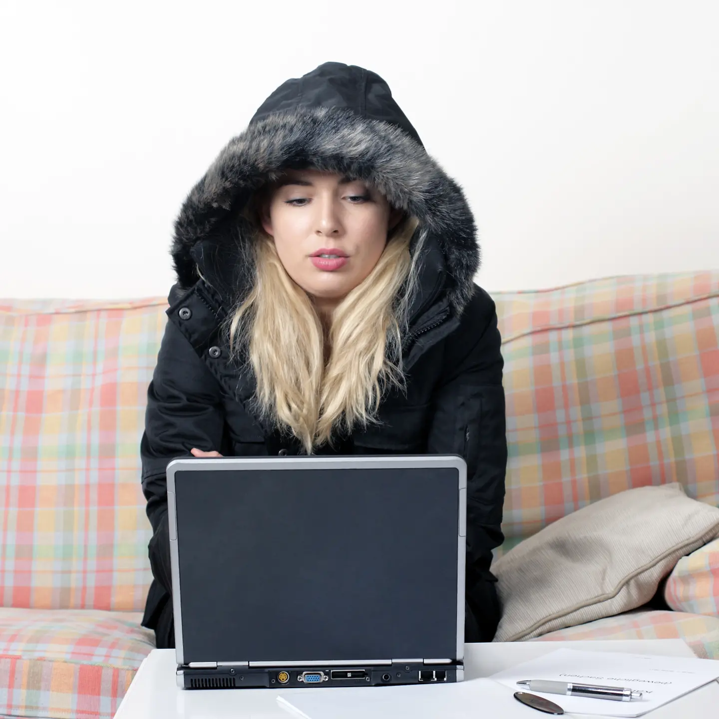 Eine Frau sitzt im waren Mantel in ihrer Wohnung auf einem Sofa vor ihrem Laptop.