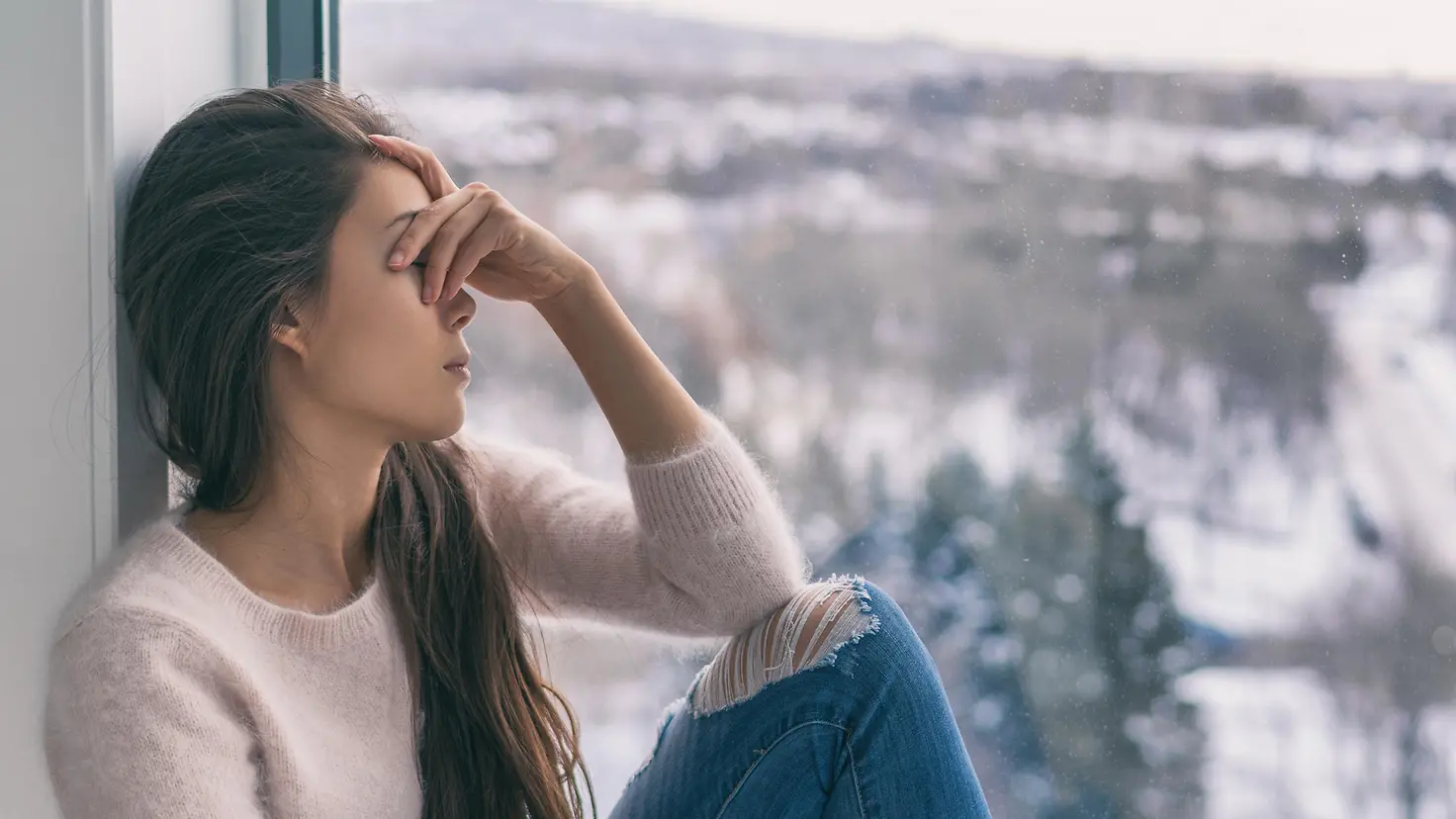 Eine junge Frau sitzt vor einem Fenster mit traurigem Blick, draußen eine Winterlandschaft