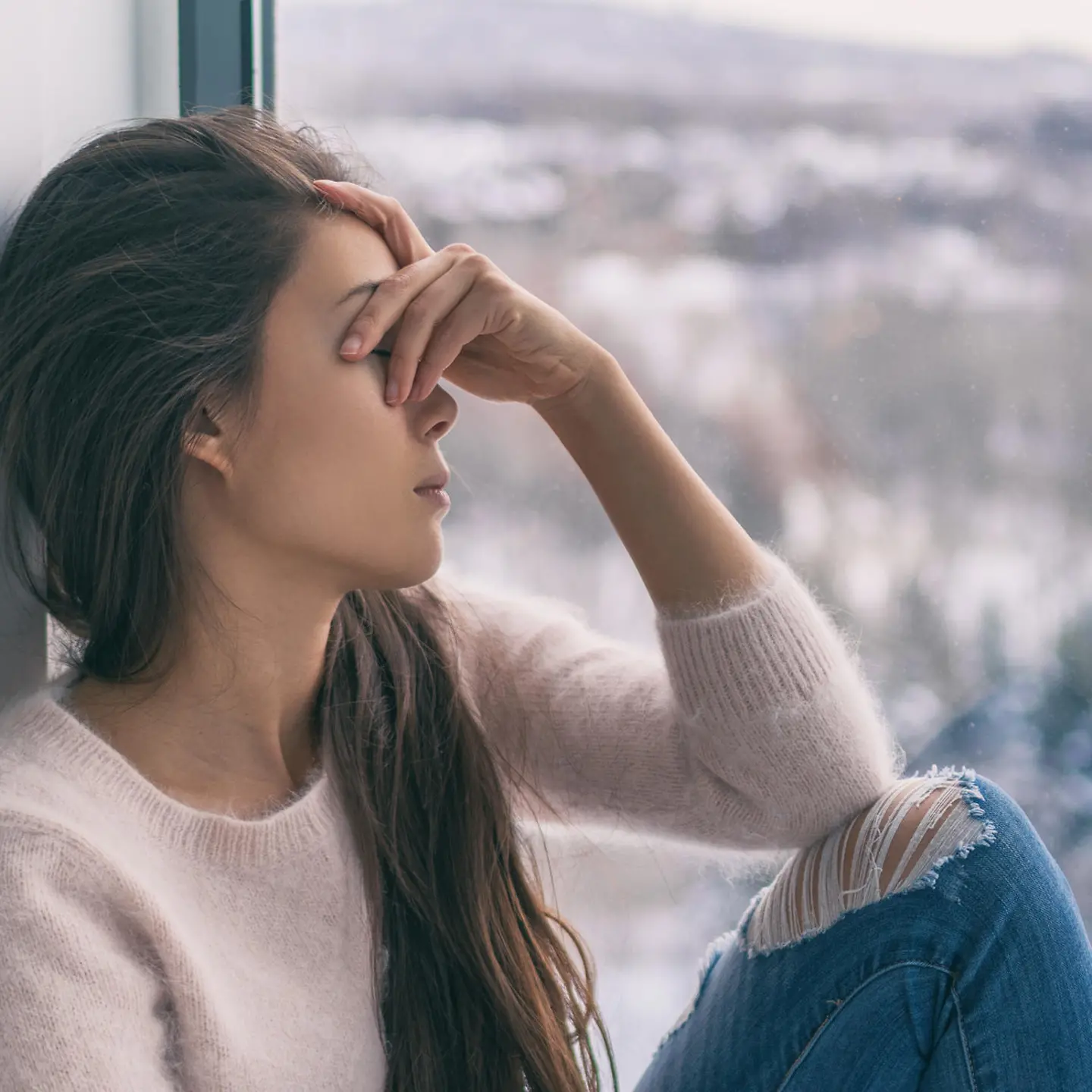 Eine junge Frau sitzt vor einem Fenster mit traurigem Blick, draußen eine Winterlandschaft
