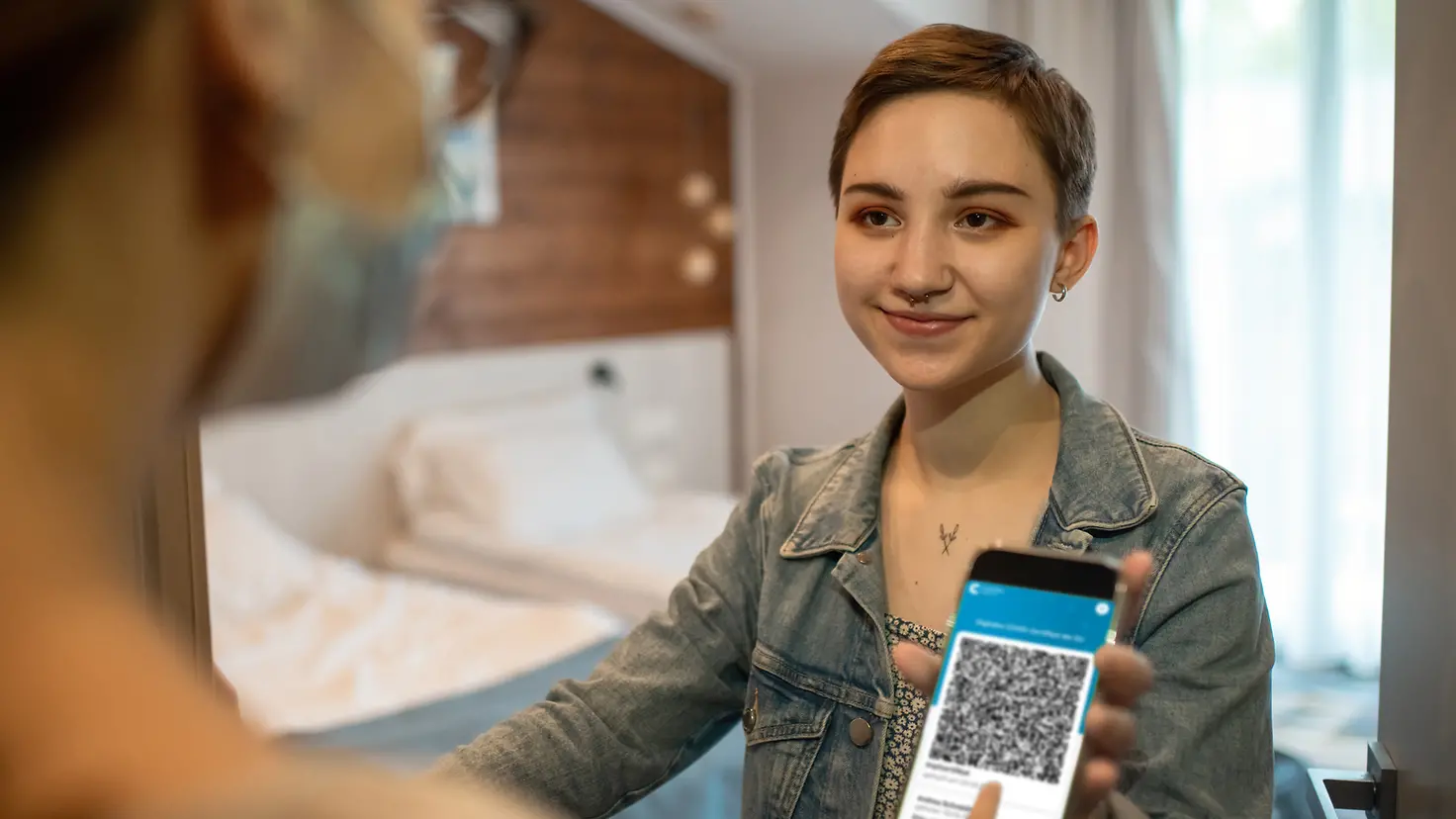 Junge Frau zeigt in einem Hotel ihren digitalen Covid-19-Impfpass mit QR-Code auf dem Smartphone.