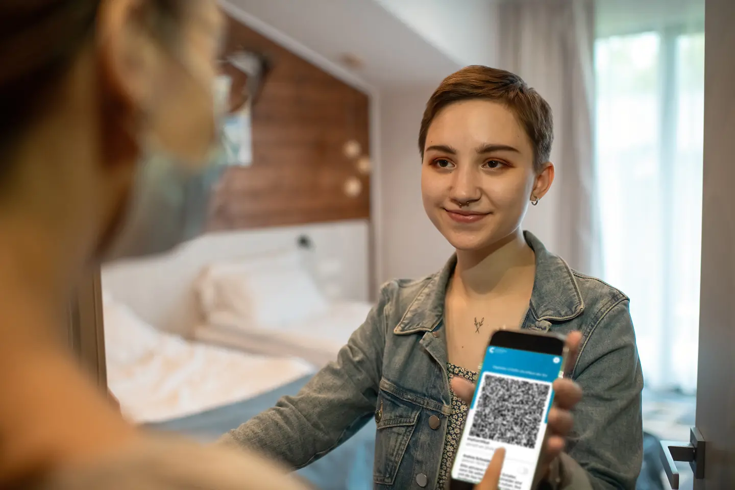 Junge Frau zeigt in einem Hotel ihren digitalen Covid-19-Impfpass mit QR-Code auf dem Smartphone.