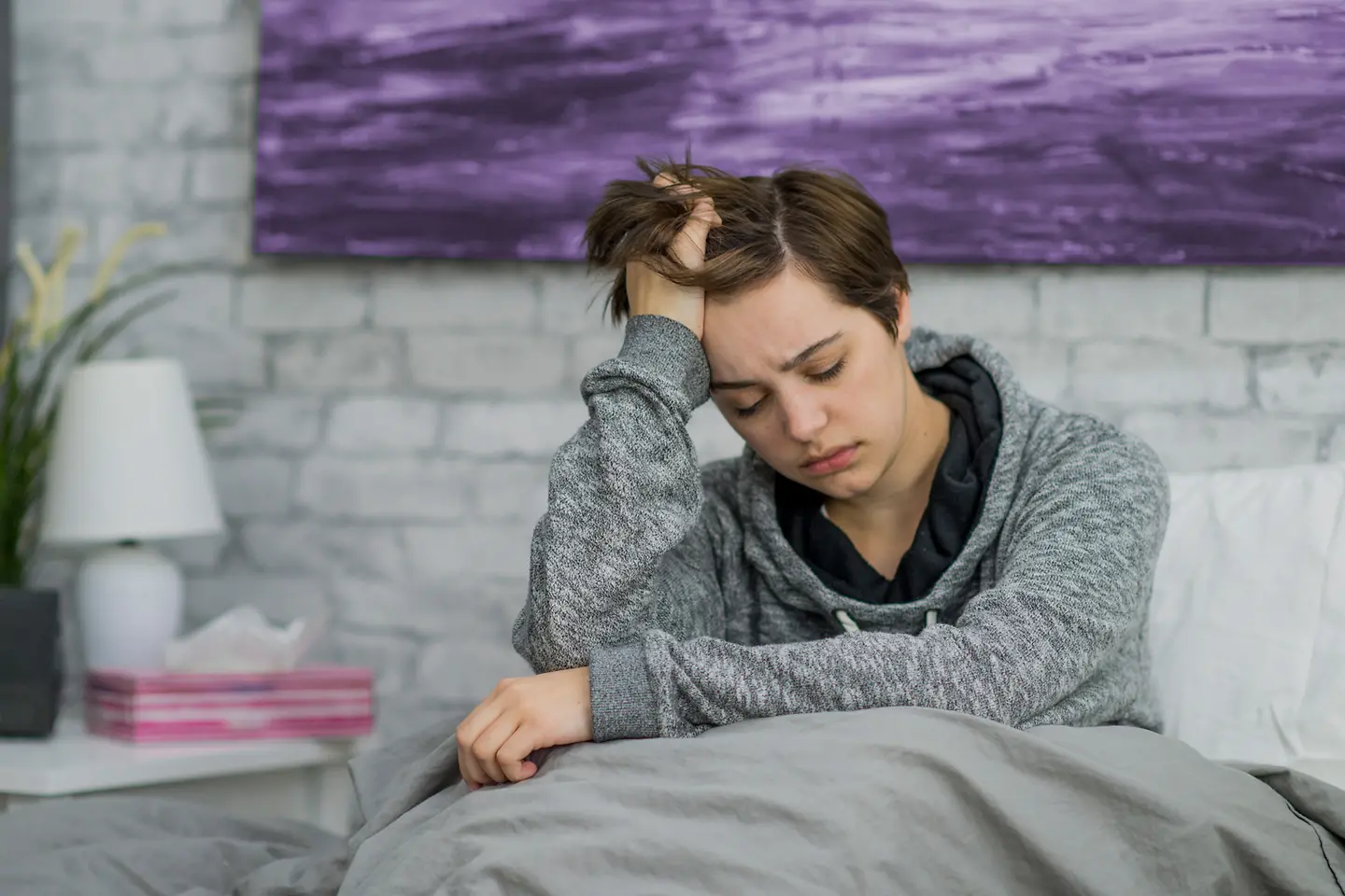 Eine junge Frau sitzt erschöpft im Bett und hält sich den Kopf; sie fühlt sich sichtbar unwohl. 