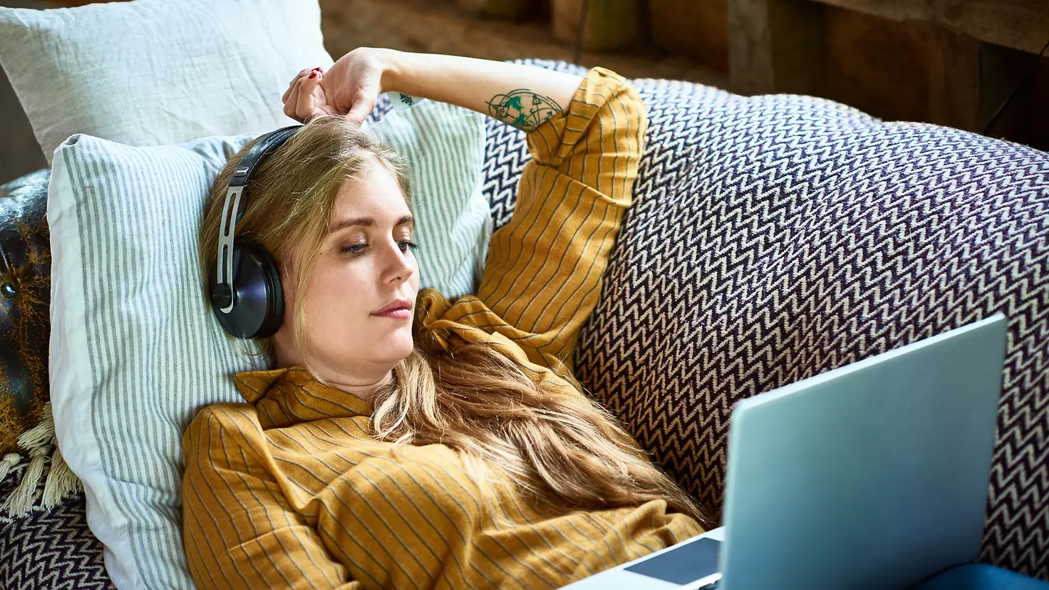 Introvertierte junge Frau hat sich mit Kopfhörern auf dem Sofa zurückgezogen, auf ihrem Schoß liegt ein aufgeklappter Laptop.