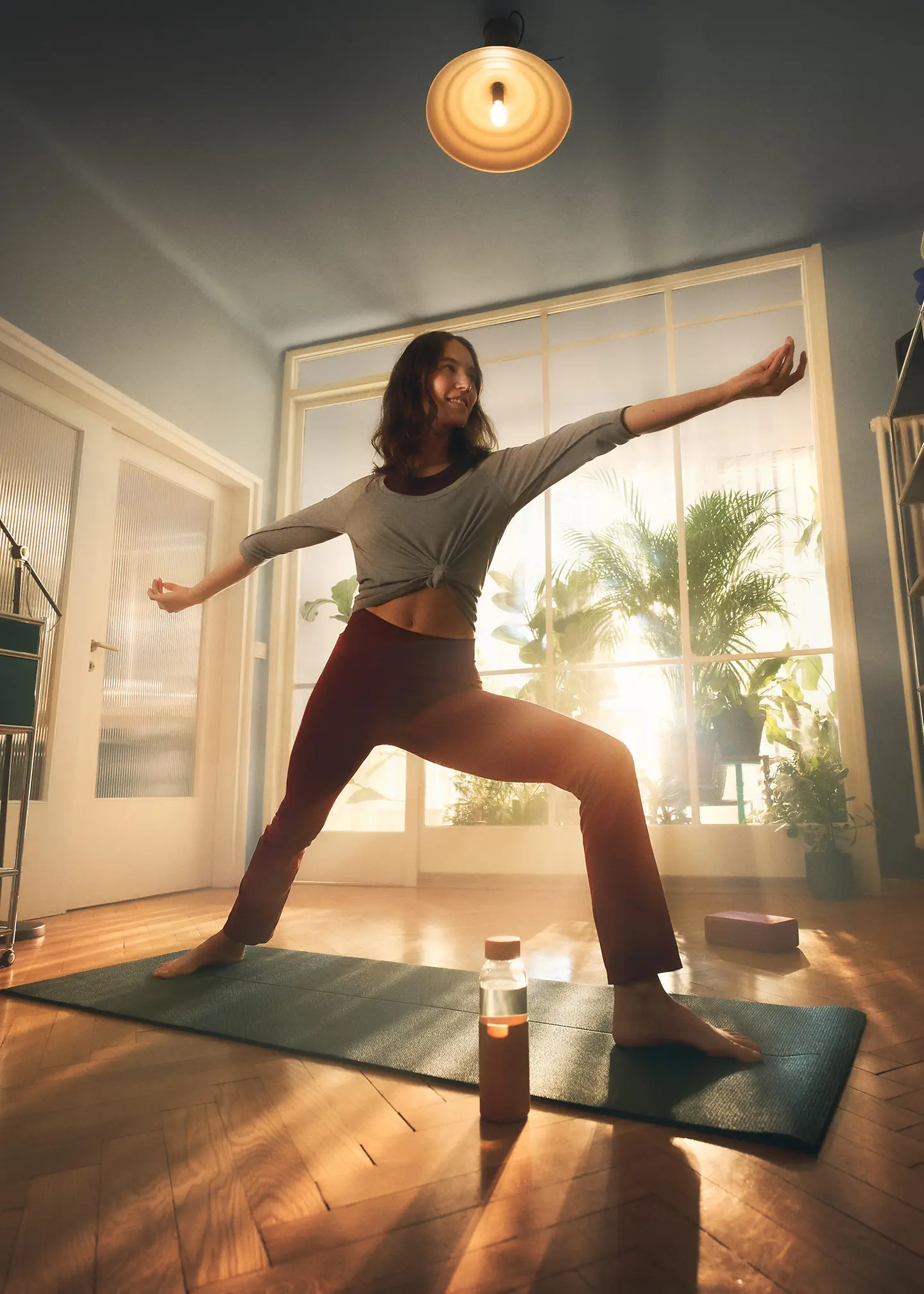 Eine Frau steht im Wohnzimmer auf einer Matte und macht eine Yogaübung.