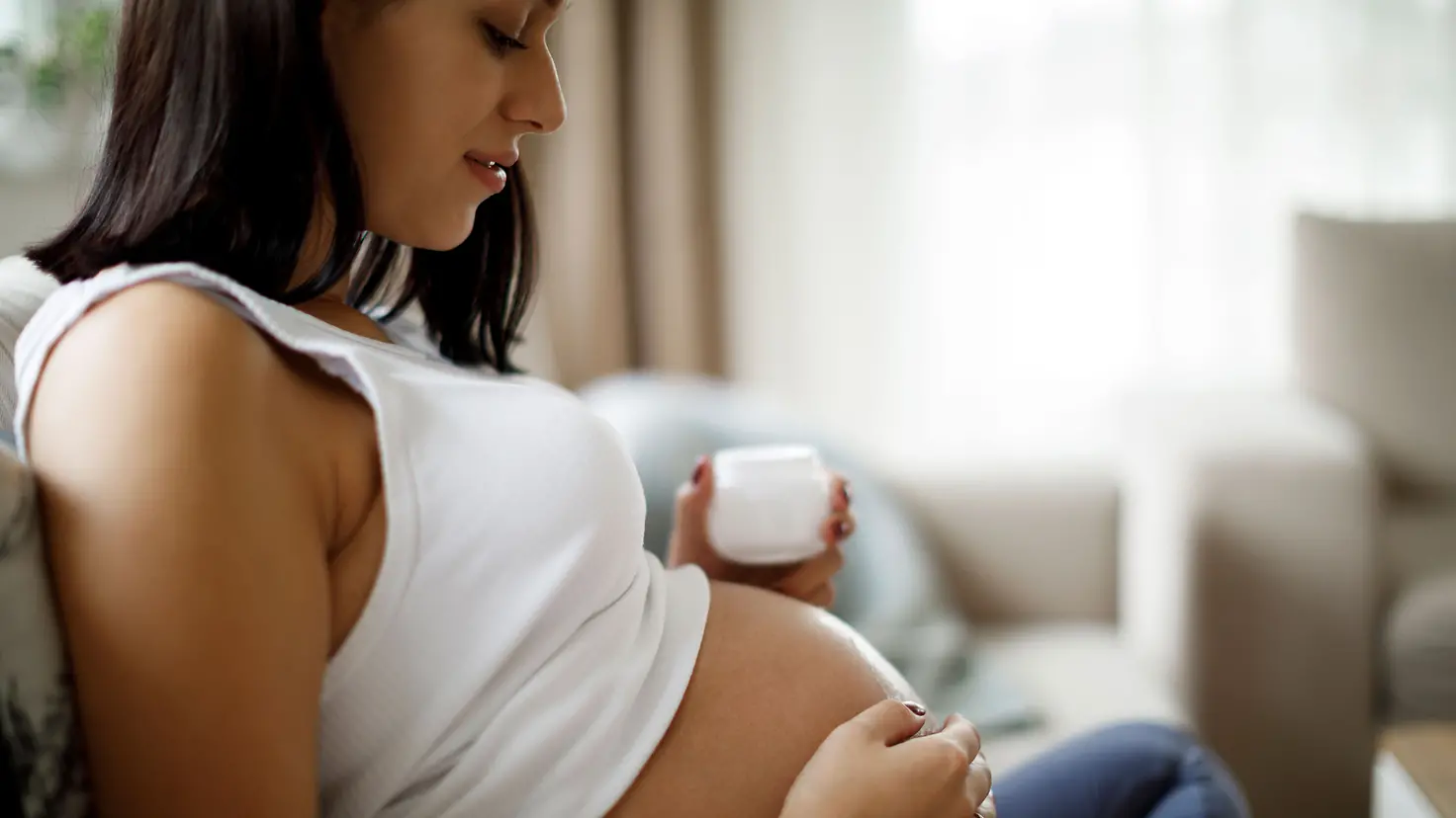 Eine schwangere Frau trägt Creme gegen Dehnungsstreifen auf ihren Babybauch auf.