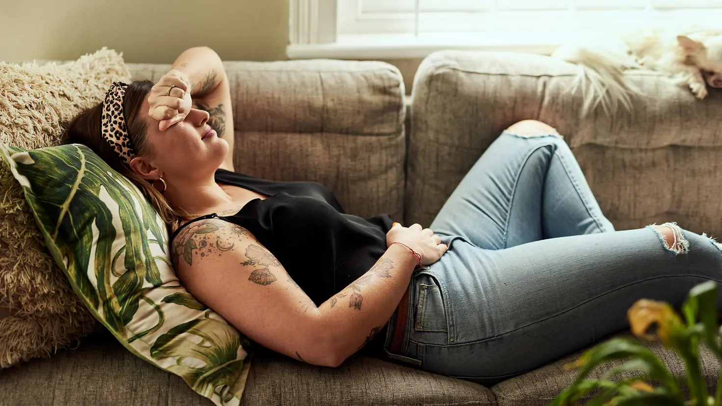 Eine junge Frau liegt aufgrund einer Eileiterschwangerschaft mit Unterleibsschmerzen auf dem Sofa. Sie hält sich eine Seite des Unterleibs und legt den anderen Arm an den Kopf.