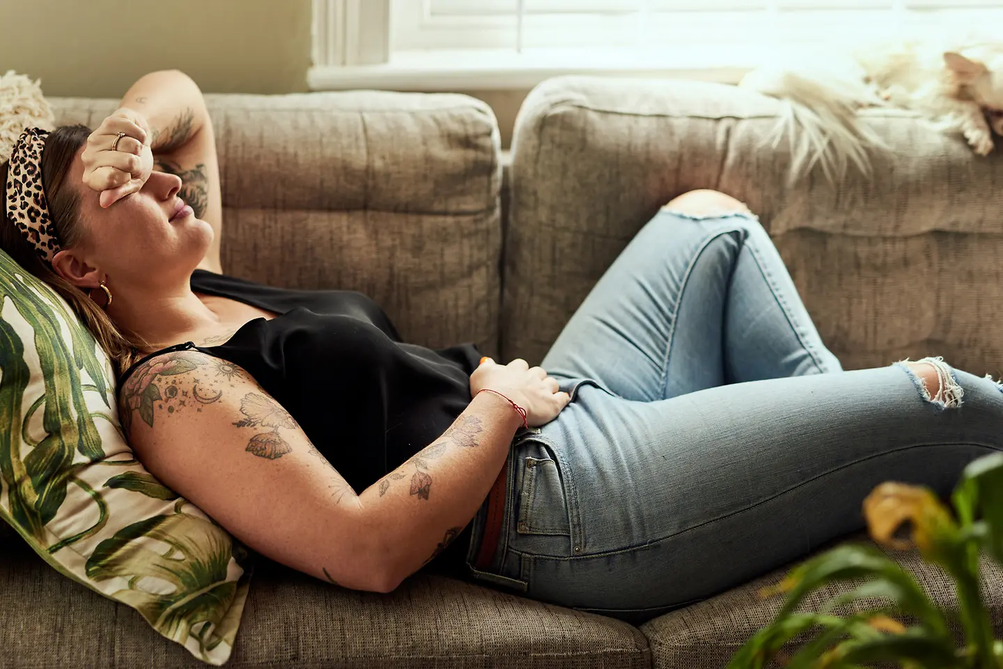 Eine junge Frau liegt aufgrund einer Eileiterschwangerschaft mit Unterleibsschmerzen auf dem Sofa. Sie hält sich eine Seite des Unterleibs und legt den anderen Arm an den Kopf.