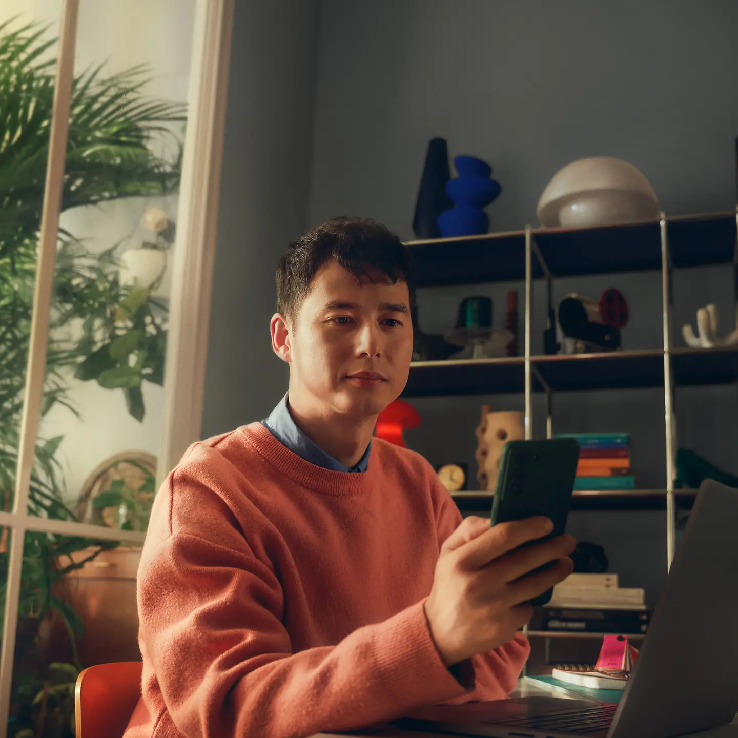 Ein Mann sitzt am Tisch vor einem Fenster und schaut auf sein Handy.