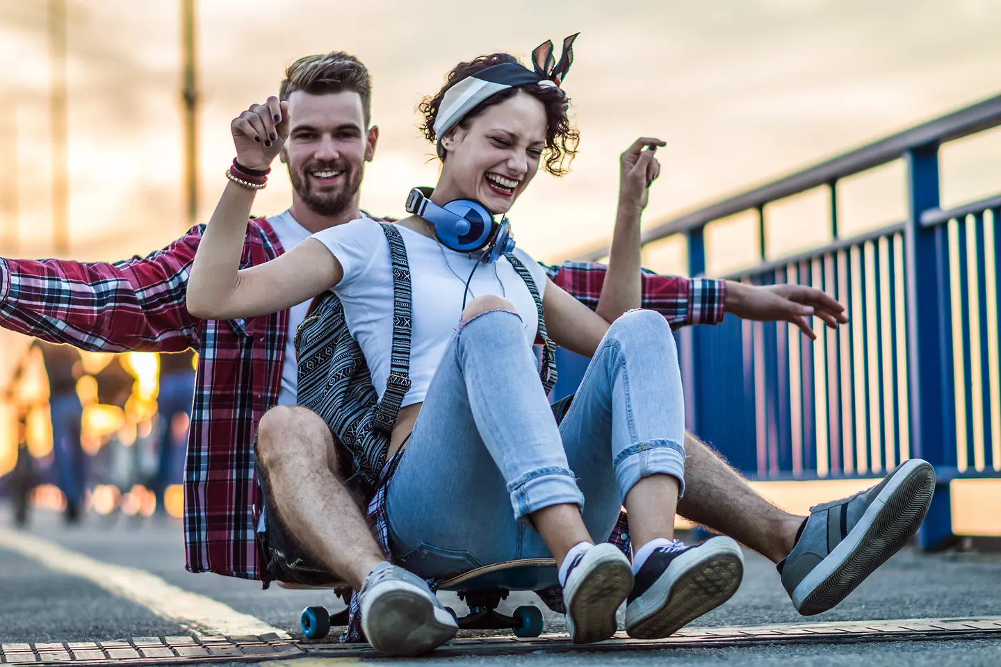 Ein junger Mann und eine extrovertierte, junge Frau sitzen lachend auf einem rollenden Skateboard.