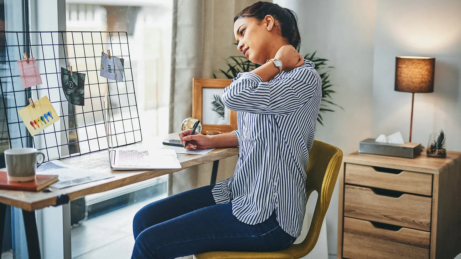 Eine Frau sitzt mit ihrem Laptop an einem kleinen Schreibtisch und hält sich den Nacken. Die Fehlhaltung durch anhaltenden Blick auf den Laptop hat zu Rückenschmerzen geführt.