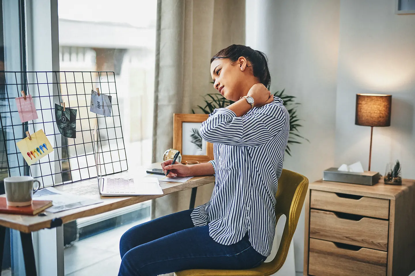 Eine Frau sitzt mit ihrem Laptop an einem kleinen Schreibtisch und hält sich den Nacken. Die Fehlhaltung durch anhaltenden Blick auf den Laptop hat zu Rückenschmerzen geführt.