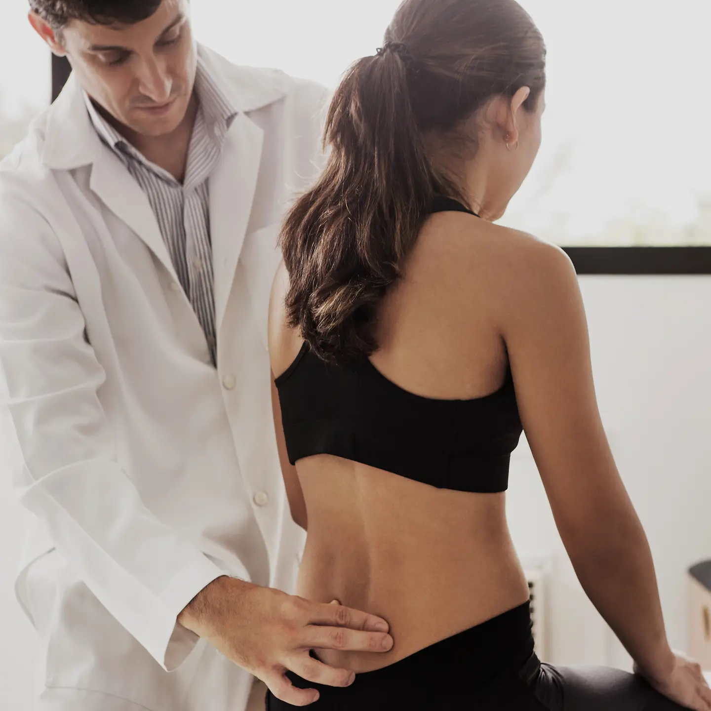 Eine junge Frau wird aufgrund ihrer starken Rückenschmerzen von einem Arzt am unteren Rücken untersucht.