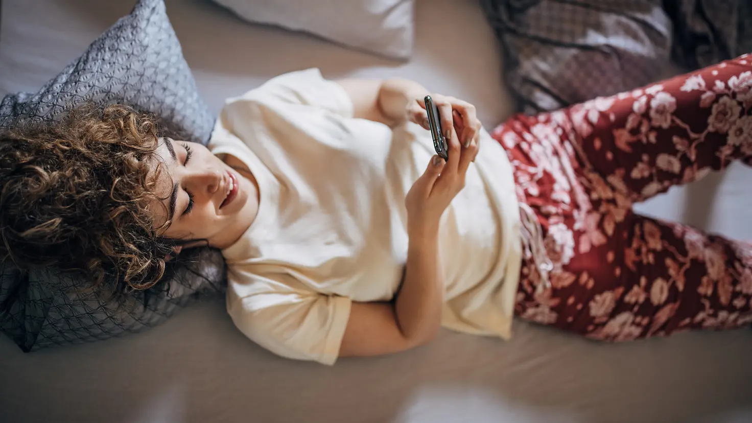 Eine junge Frau im Bett verfolgt über die Barmer-App auf ihrem Mobiltelefon den Status ihrer Krankmeldung.