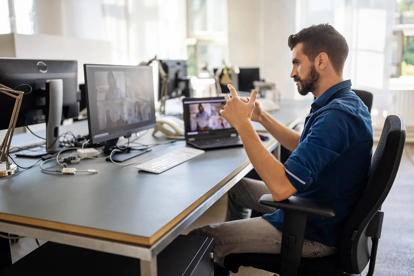 Ein junger Mann sitzt am Schreibtisch mit Monitor und Laptop. Sein Schreibtischstuhl ist an die Anatomie der Wirbelsäule angepasst.