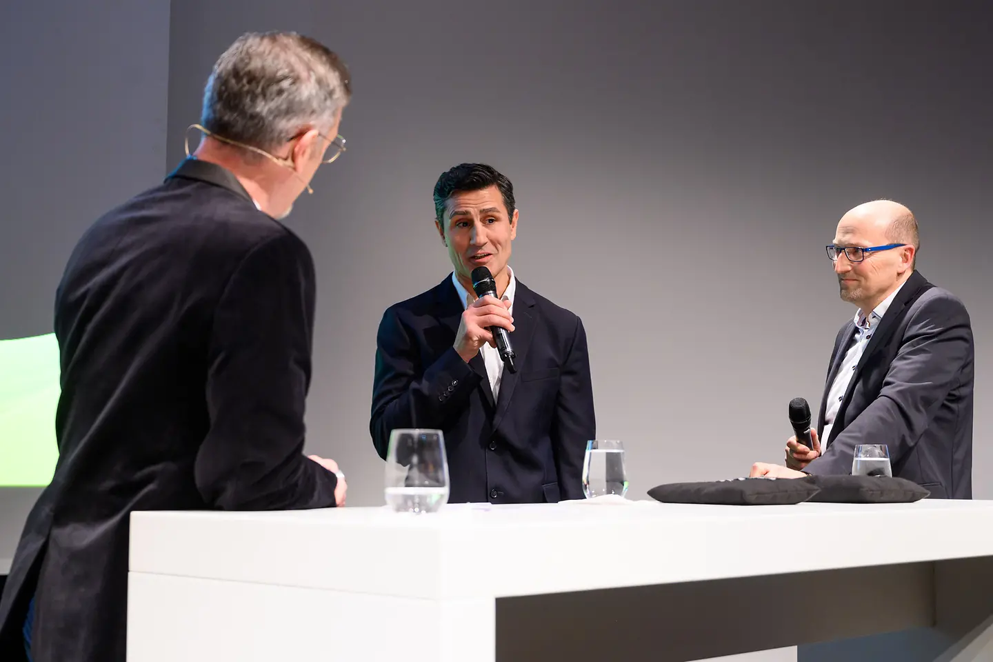 Ein Mann hält ein Mikrofon in der Hand und redet, zwei andere Männer hören ihm zu