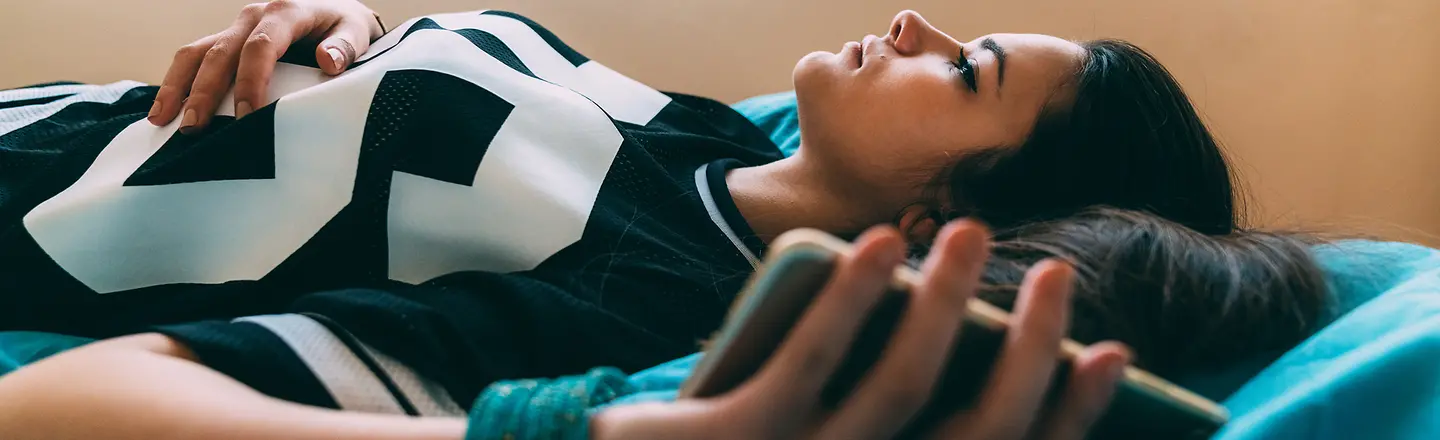 Eine niedergeschlagene junge Frau liegt mit dem Smartphone in der Hand auf dem Bett. Sie ist Opfer von Cybermobbing geworden.