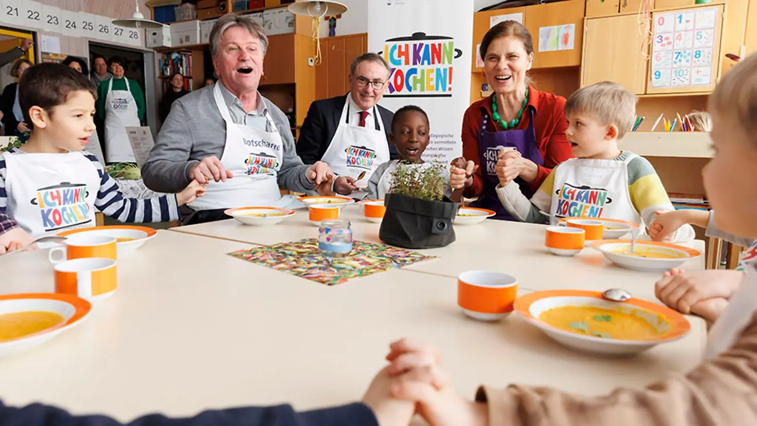 Der baden-württembergische Gesundheitsminister Manne Lucha, der Barmer-Vorstandsvorsitzende Professor Dr. Christoph Straub und die Köchin Sarah Wiener sitzen mit Kindern an einem Tisch und halten sich an den Händen.