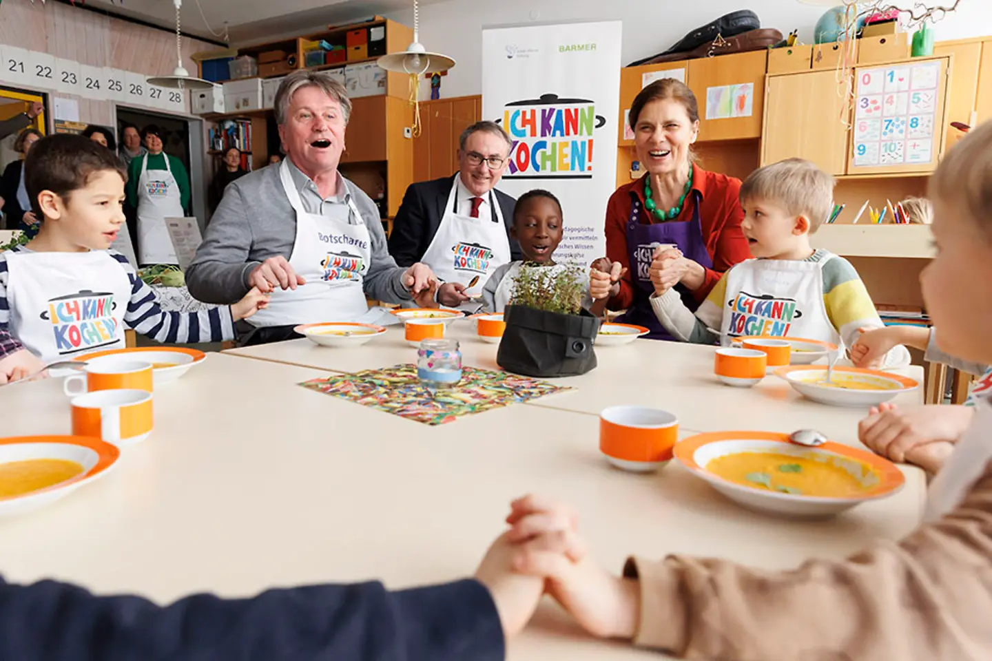Der baden-württembergische Gesundheitsminister Manne Lucha, der Barmer-Vorstandsvorsitzende Professor Dr. Christoph Straub und die Köchin Sarah Wiener sitzen mit Kindern an einem Tisch und halten sich an den Händen.