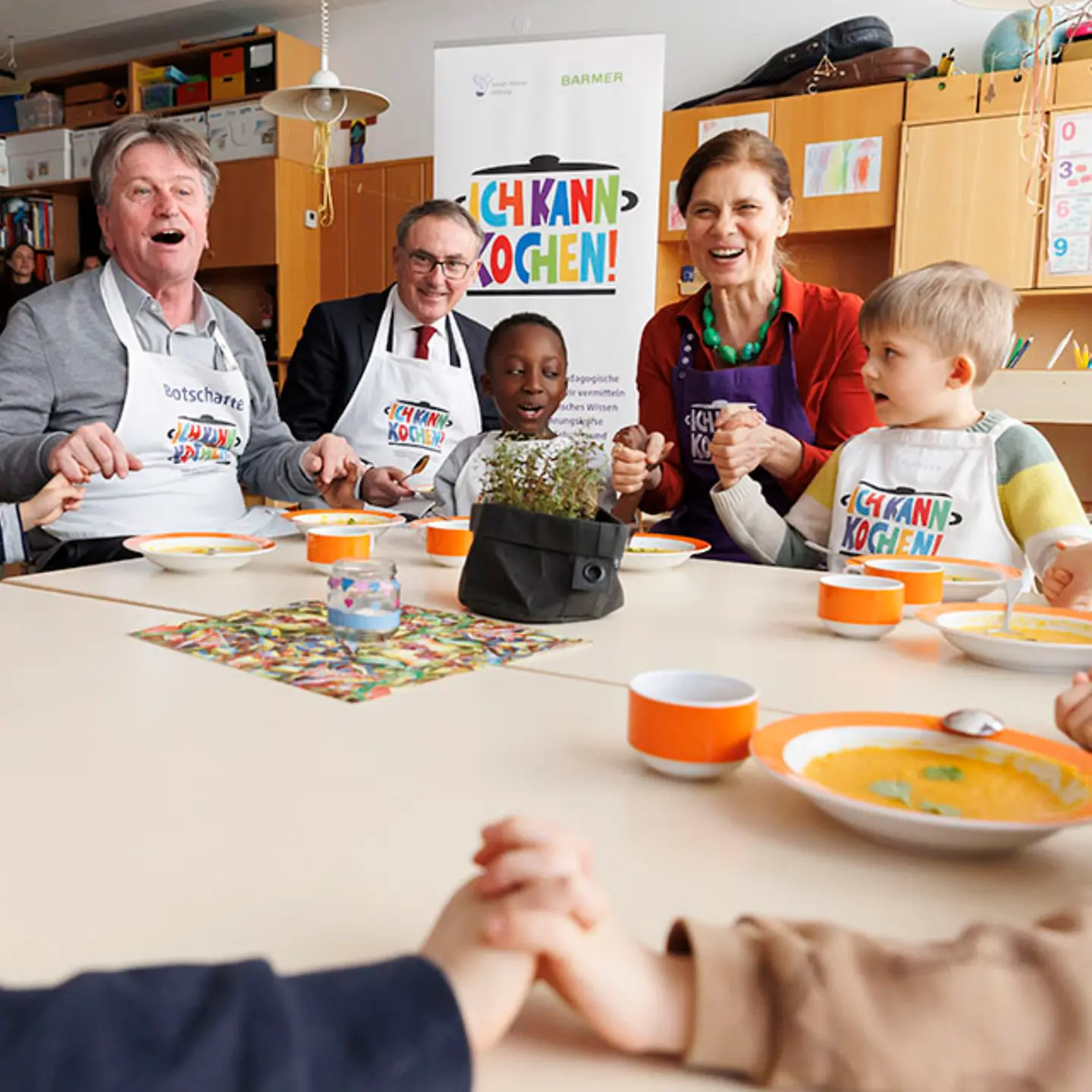 Der baden-württembergische Gesundheitsminister Manne Lucha, der Barmer-Vorstandsvorsitzende Professor Dr. Christoph Straub und die Köchin Sarah Wiener sitzen mit Kindern an einem Tisch und halten sich an den Händen.