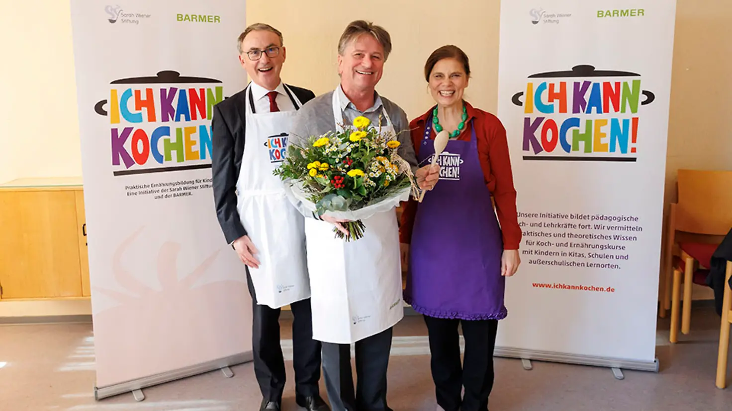 Barmer-Vorstandsvorsitzender Prof. Dr. Christoph Straub, Manne Lucha mit einem Blumenstrauß in der Hand und Köchin Sarah Wiener stehen zusammen und lächeln in die Kamera.