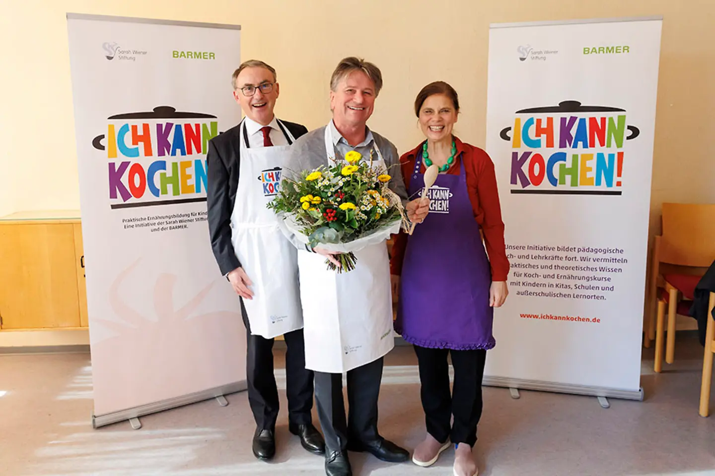 Barmer-Vorstandsvorsitzender Prof. Dr. Christoph Straub, Manne Lucha mit einem Blumenstrauß in der Hand und Köchin Sarah Wiener stehen zusammen und lächeln in die Kamera.