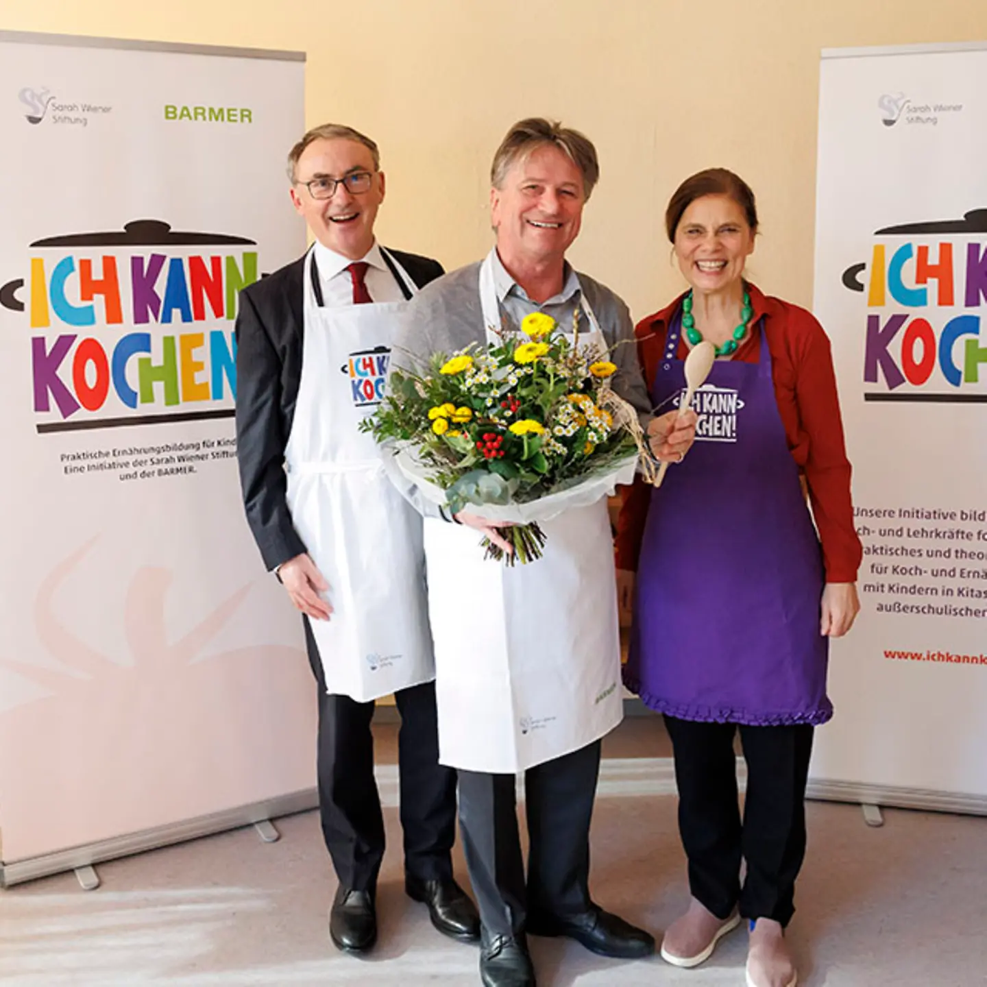 Barmer-Vorstandsvorsitzender Prof. Dr. Christoph Straub, Manne Lucha mit einem Blumenstrauß in der Hand und Köchin Sarah Wiener stehen zusammen und lächeln in die Kamera.