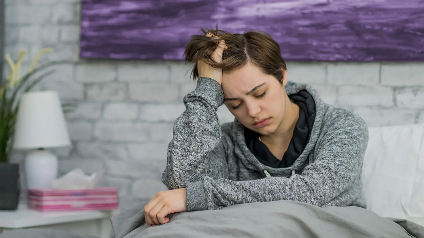 Eine junge Frau sitzt im Bett und leidet unter Einsamkeit und Kopfschmerzen, sie fühlt sich sichtbar unwohl. 