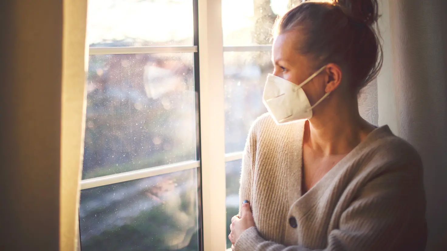 Eine einsame Frau mit FFP2-Maske steht am Fenster.