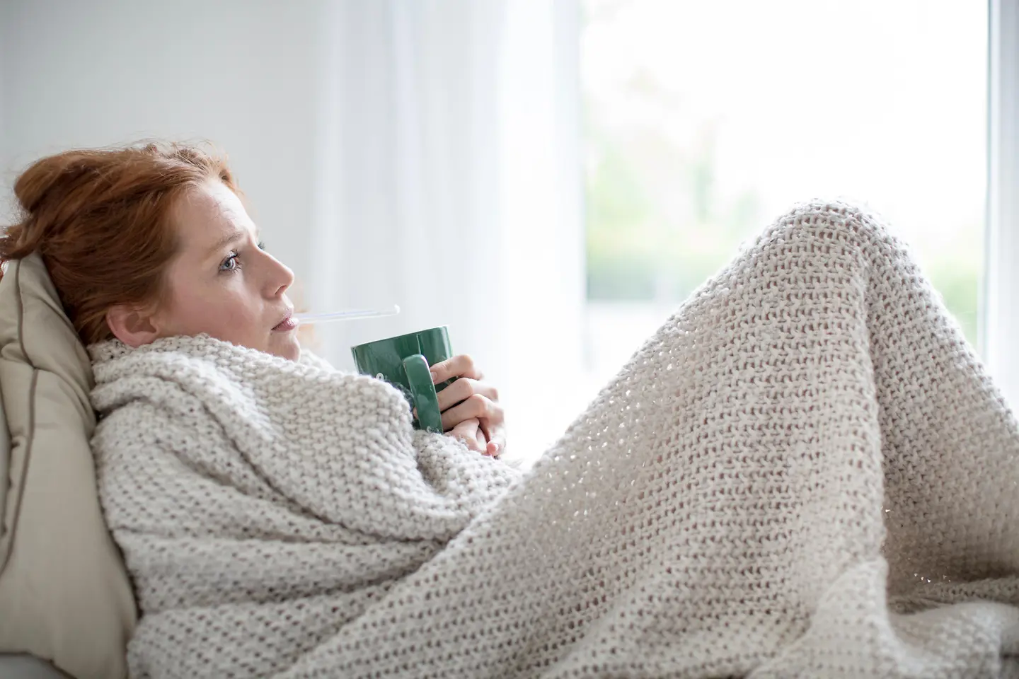 Zu sehen ist eine sitzende Frau eingehüllt in Decke mit Tasse in der Hand 