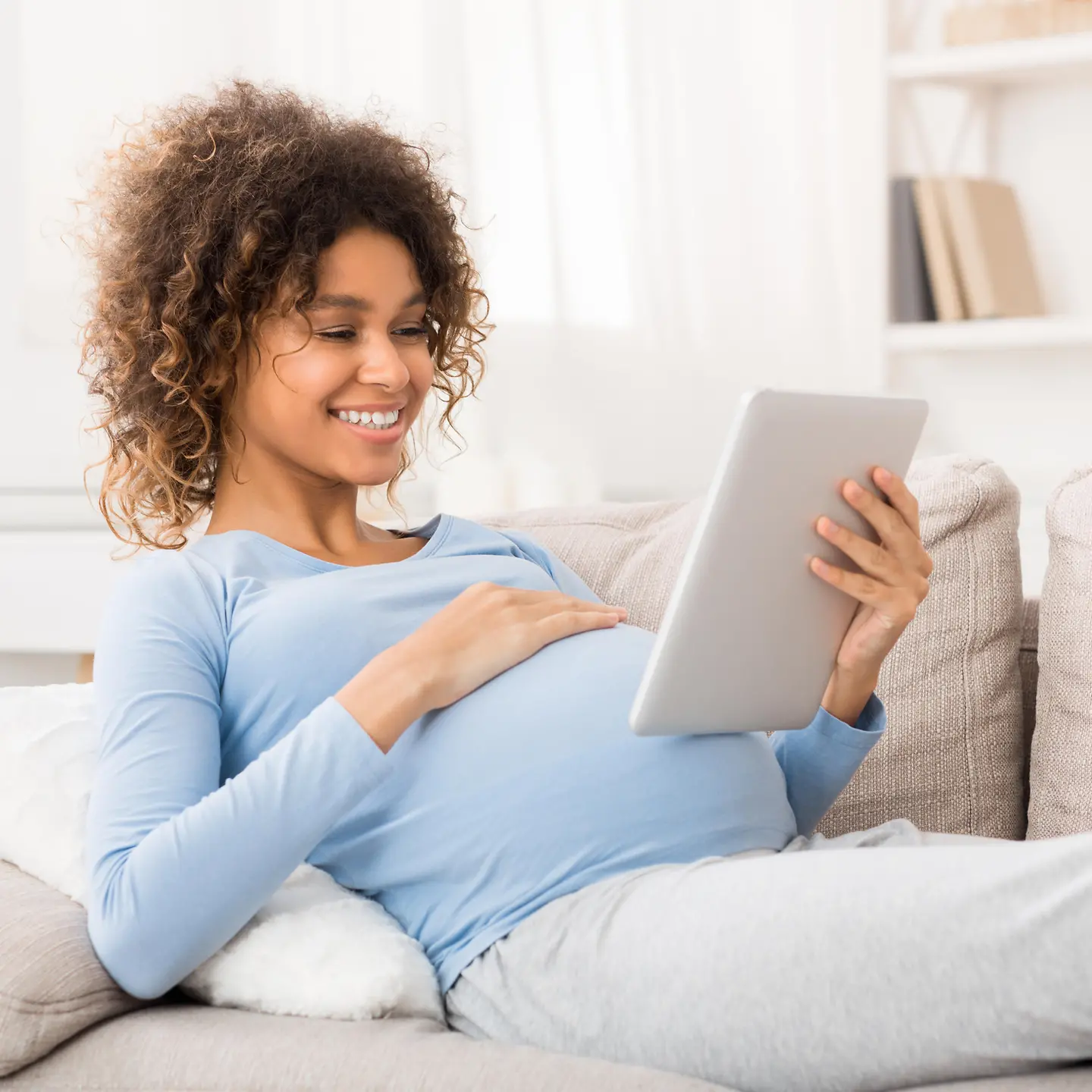 Eine schwangere Frau liegt auf dem Sofa und schaut auf ein Tablet, das sie in ihrer Hand hält.