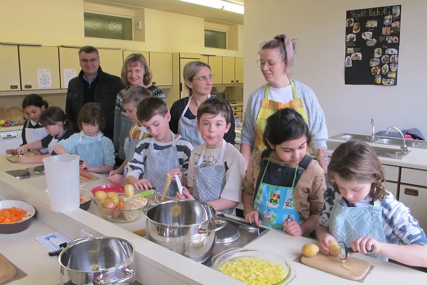 Kinder schnippeln Gemüse an einer langen Küchenarbeitsfläche, dahinter die Genussbotschafterinnen, BARMER-Geschäftsführer Zöhler und Schulleitung.