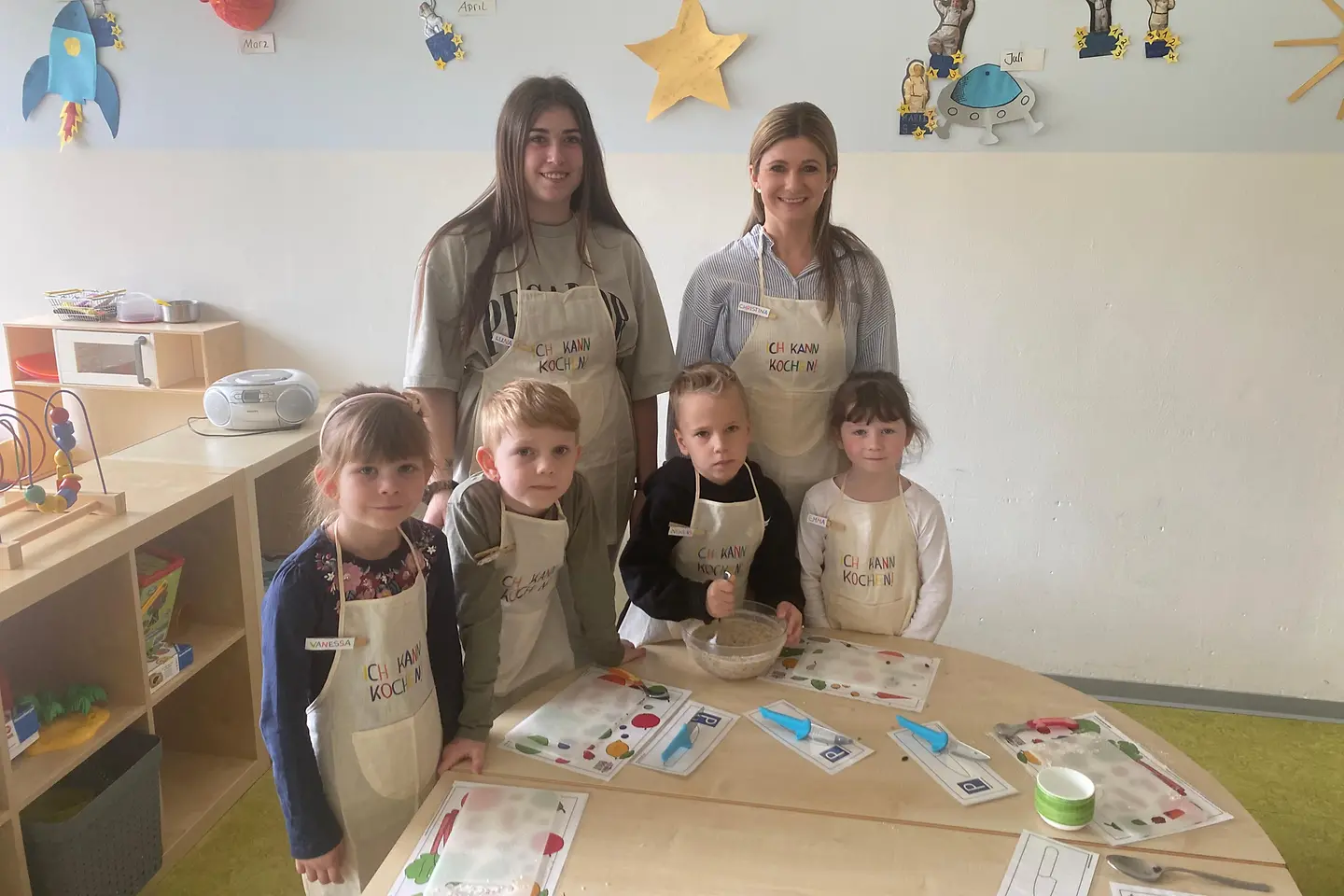Kinder stehen mit zwei Erzieherinnen am Tisch und bereiten Essen zu.