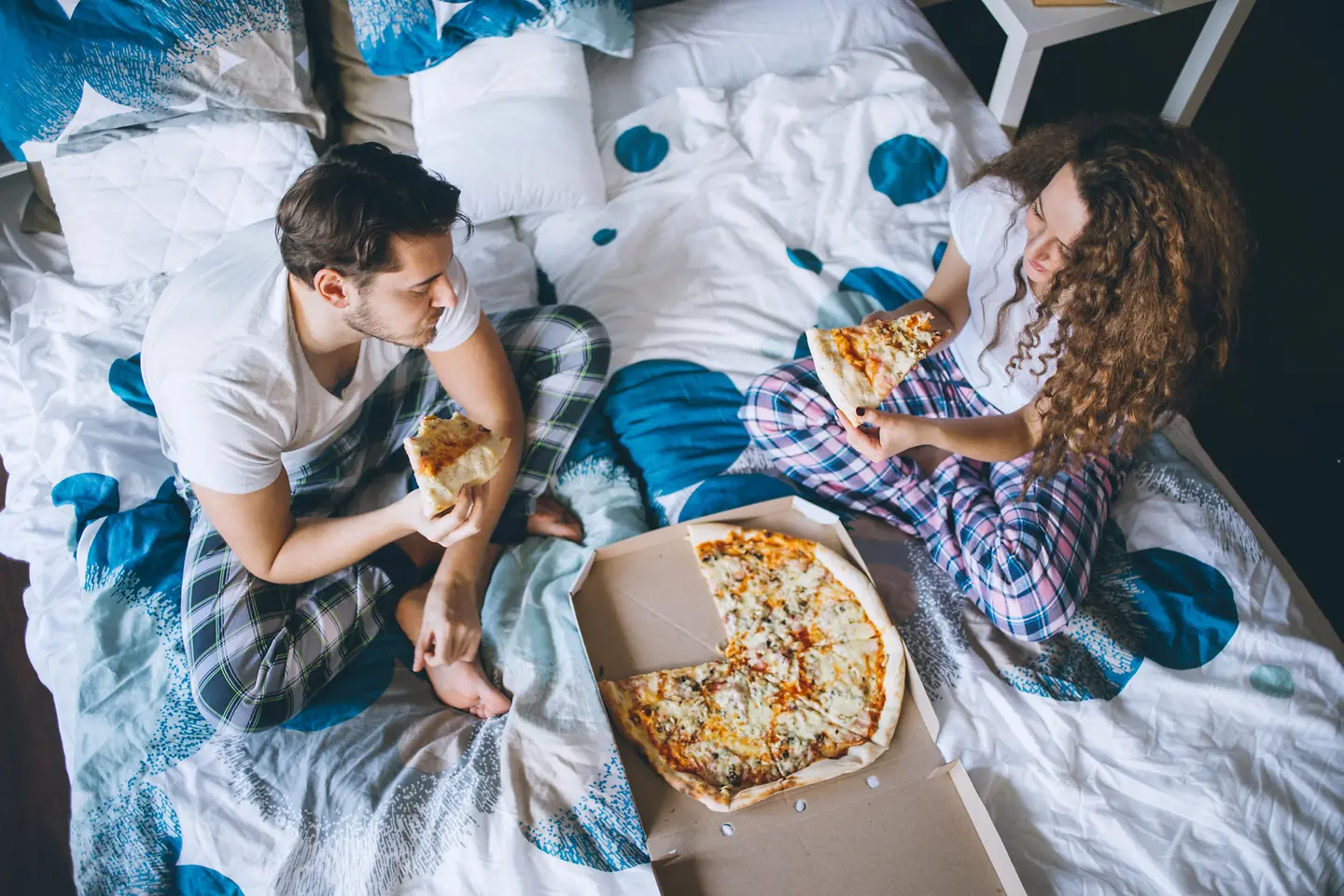Zwei Personen sitzen auf dem Bett und essen genüsslich eine Pizza
