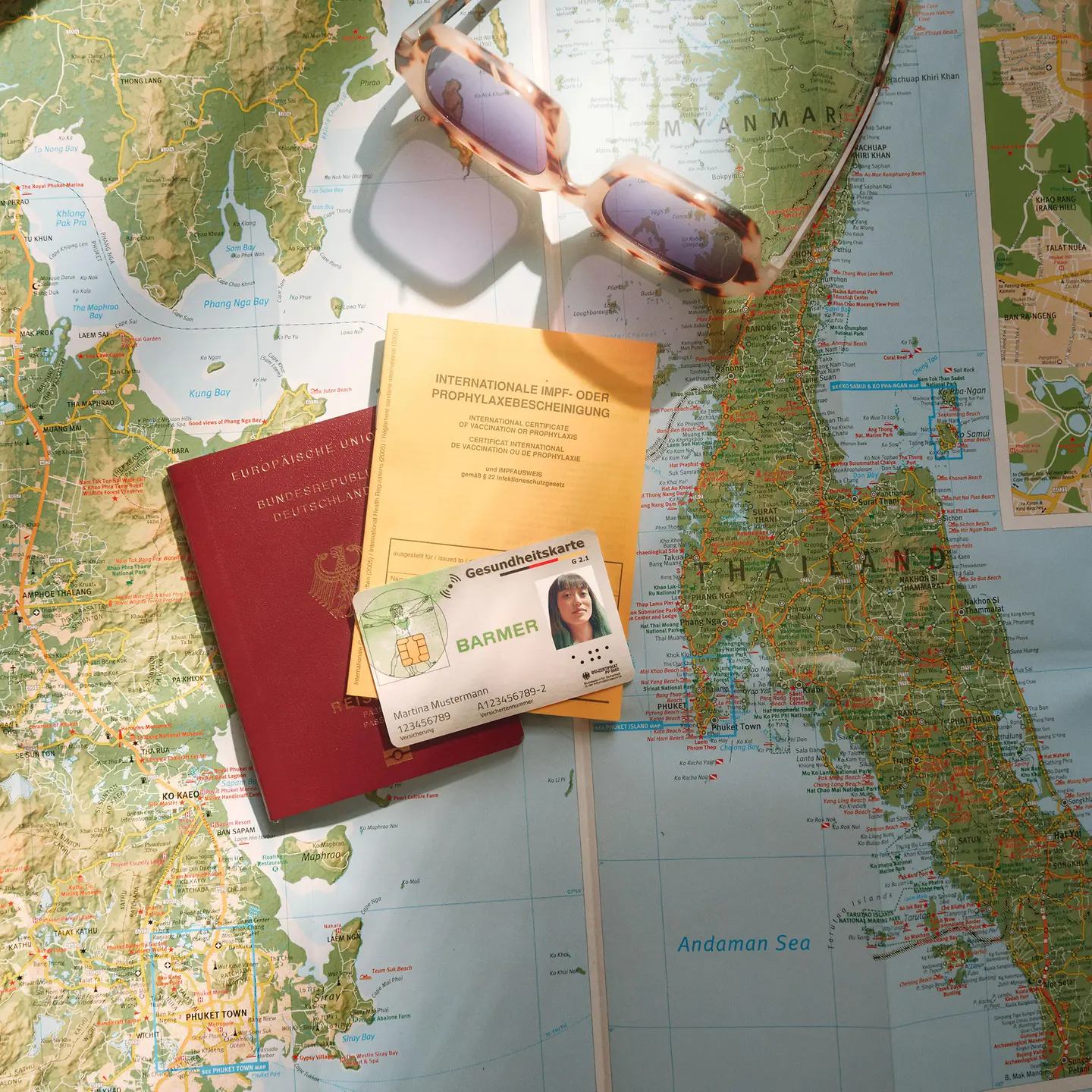 Auf einer ausgefalteten Weltkarte liegen eine Barmer-Versichertenkarte, ein Reisepass, ein Impfausweis und eine Sonnenbrille. 