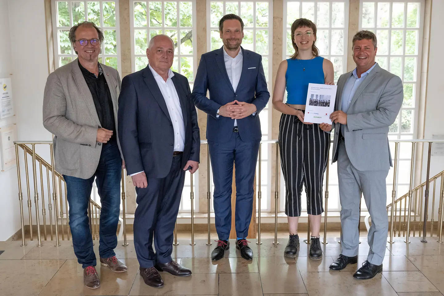 Fünf Projektverantwortliche stehen vor einem Geländer und halten den Abschlussbericht zu Healthy Campus Mainz in die Kamera.