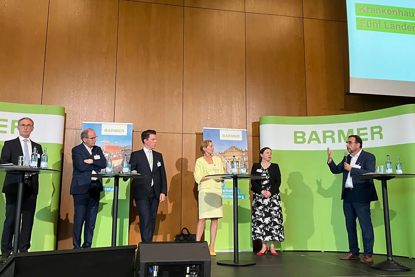 Podiumsdiskussion, ein Mann hält ein Mikrofon in seiner Hand und redet, die anderen Teilnehmer hören zu.