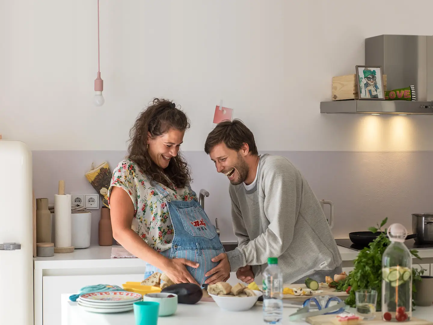 Eine schwangere Frau und ein Mann stehen in der Küche und lachen