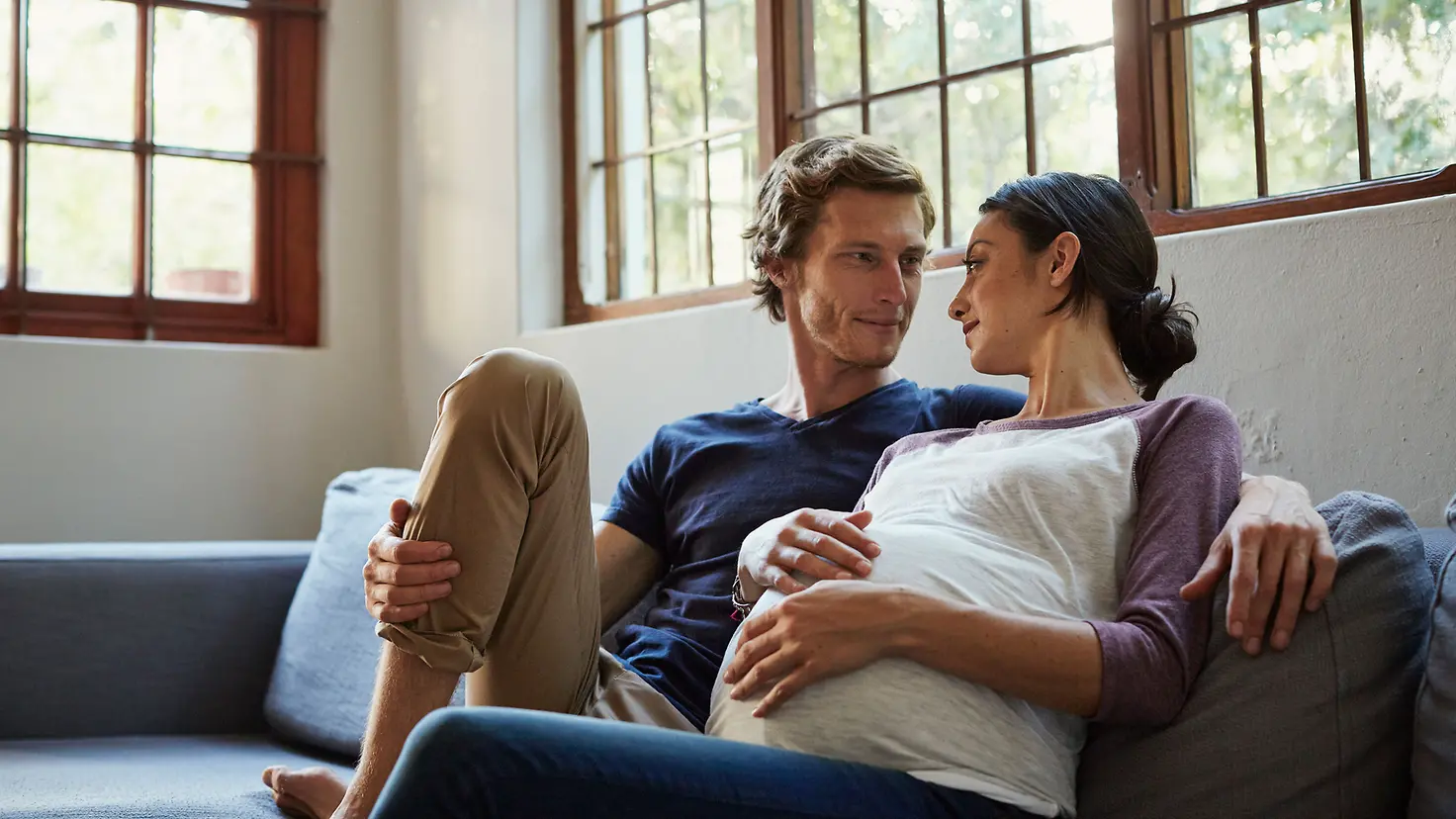 Eine hochschwangere Frau und ihr Partner schauen sich an, während sie sich auf dem Sofa entspannen