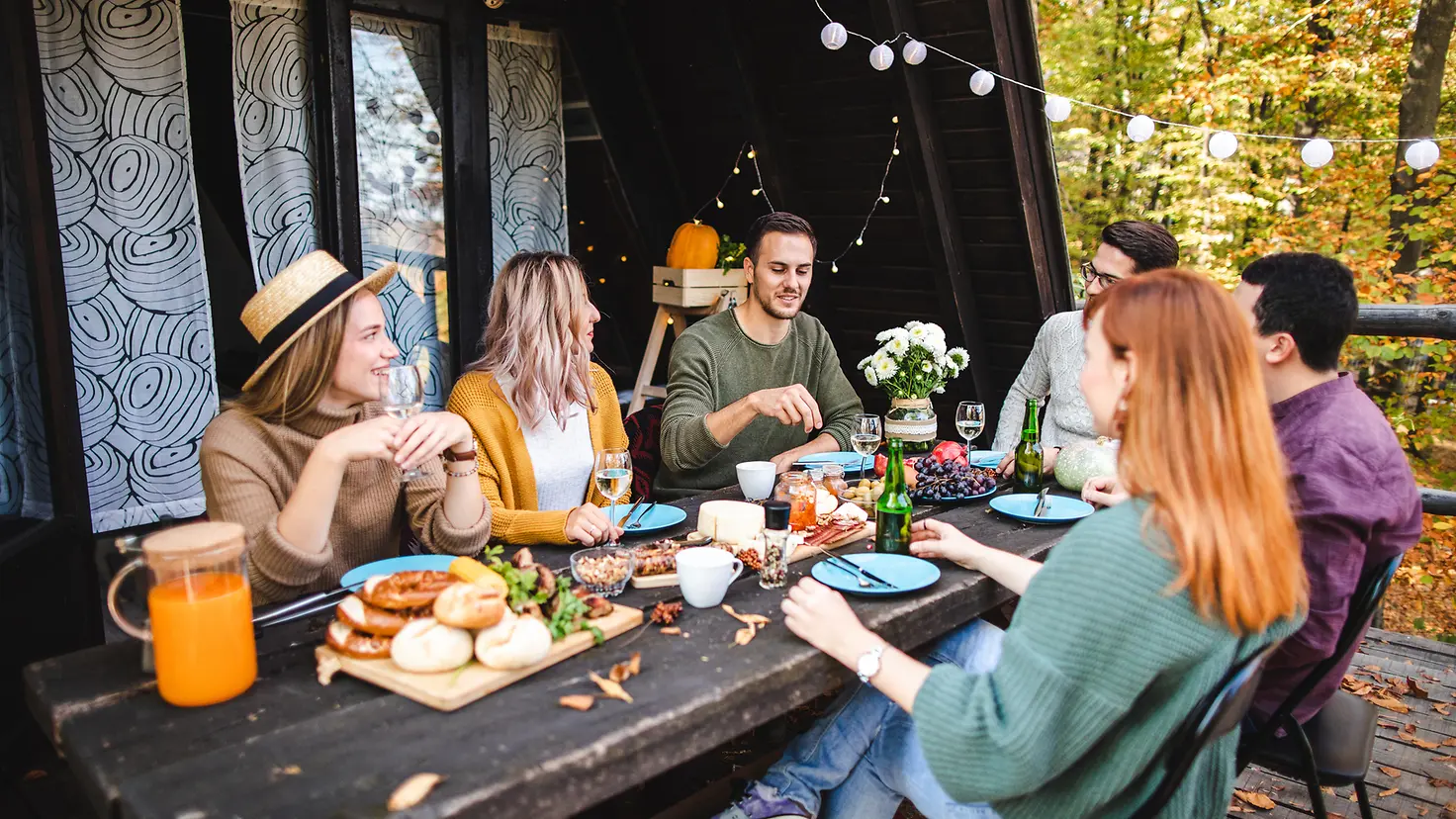 Junge Frauen und Männer sitzen an einem Tisch vor einem Holzhaus und essen gemeinsam..