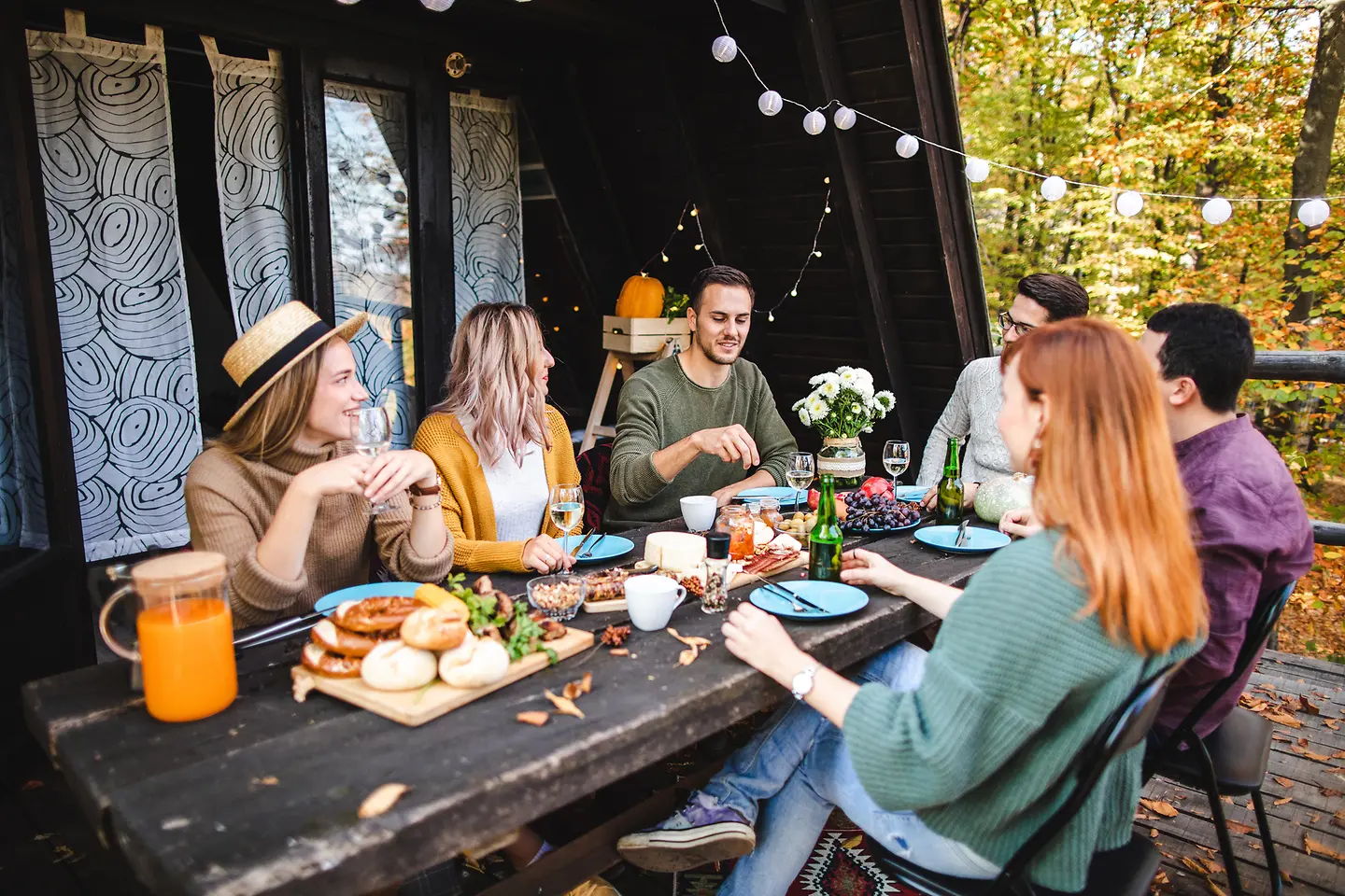 Junge Frauen und Männer sitzen an einem Tisch vor einem Holzhaus und essen gemeinsam..